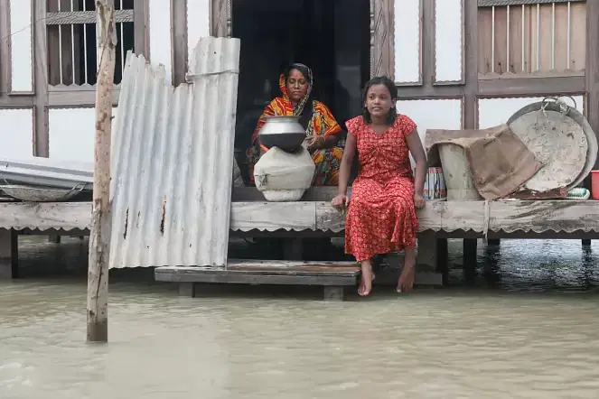 A woman cooks and a girl looks ahead sitting on a house sit in floodwater at Medinimandal, Munshiganj on 7 July 2020.