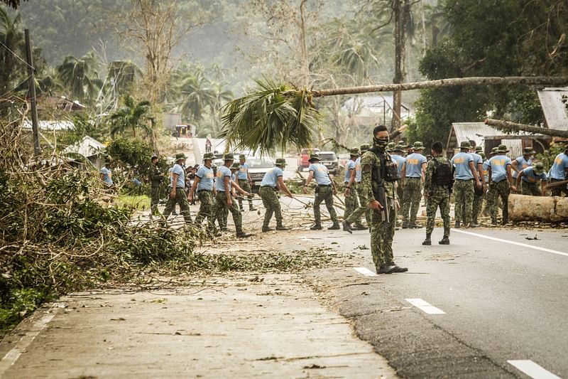 An armed policeman stands guard while his colleagues clear a road of debris in Surigao City, Surigao del norte province, on 19 December, 2021, days after super Typhoon Rai devastated the city