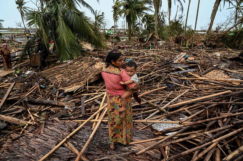 A Rohingya woman carries her baby next to her destroyed house at Basara refugee camp in Sittwe on 16 May, 2023, after Cyclone Mocha made a landfall. The death toll from Cyclone Mocha that barrelled through Myanmar has reached 145 in the country, the junta's information team said in a statement on 19 May.