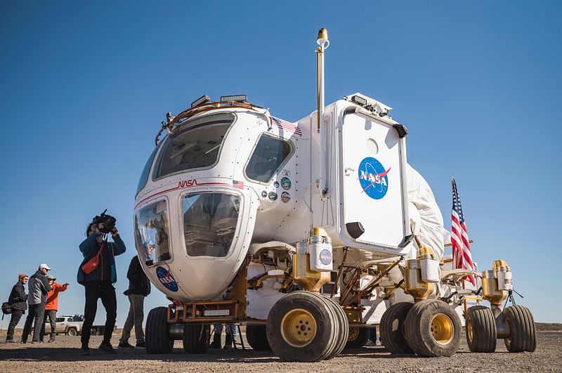 In this file photo taken on 24 October, 2022 members of the media stand next to a Moon rover prototype for future Artemis missions at the Black Point Lava Flow near Flagstaff, Arizona. The next time NASA goes to the Moon, it intends to stay. Under the Artemis programme, the US space agency plans to colonize, for the very first time, a celestial body other than Earth. But building a lunar base is no simple feat.