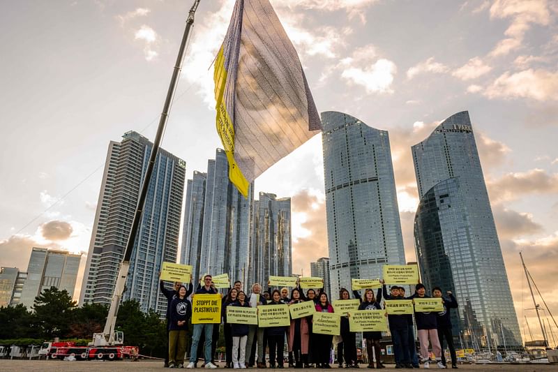 A flag with an eye and writing that reads “Governments, the world is watching, cut plastic production now” is displayed above Greenpeace activists holding placards in Busan on November 25, 2024, before the opening of the Fifth session of UN Intergovernmental Negotiating Committee on Plastic Pollution (INC-5) at a nearby venue.