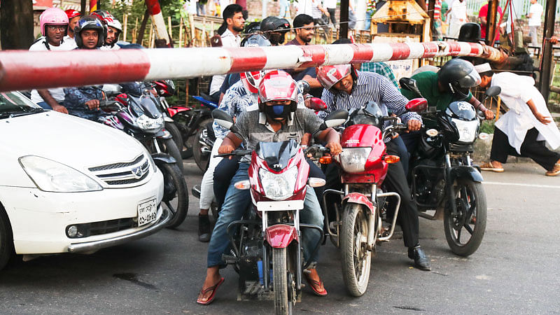 ট্রেন আসার আগ মুহূর্তে যান চলাচল বন্ধ করতে ফেলা হয়েছে প্রতিবন্ধক। সেটির নিচ দিয়ে কসরত করে বের হয়ে যাওয়ার চেষ্টা করছেন মোটরসাইকেলের চালকেরা। সেকেন্ডের ব্যবধানে ঘটে যেতে পারে বড় দুর্ঘটনা। গতকাল রাজধানীর কারওয়ান বাজারে এফডিসি রেলক্রসিং এলাকায়।  ছবি: প্রথম আলো