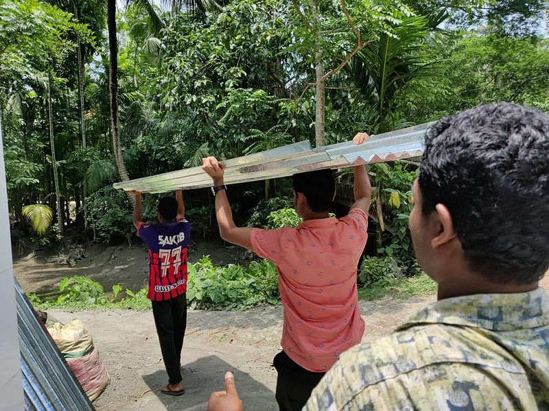 বৃদ্ধা হাজেরা বেগমের ঘর তৈরির জন্য ঢেউটিন নিয়ে যাচ্ছেন স্বেচ্ছাসেবকেরা