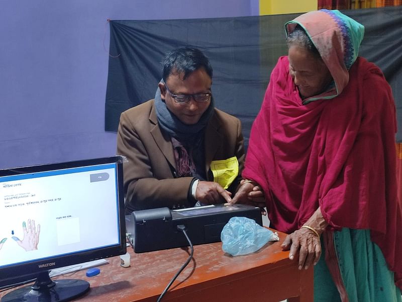 ইভিএম মেশিনে আঙুলের ছাপ মেলাতে ভোটারদের সহায়তা করছেন সহকারী প্রিসাইডিং কর্মকর্তা