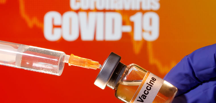 A small bottle labeled with a "Vaccine" sticker is held near a medical syringe in front of displayed "Coronavirus COVID-19" words in this illustration 