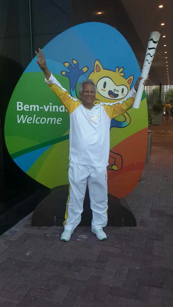 Nobel laureate professor muhammad Yunus holding up the Olympic Torch in one hand and displaying three fingers with another hand to symbolize three zeros in front the picture of Rio Olympic 2026 Mascot. Photo: Yunus Centre