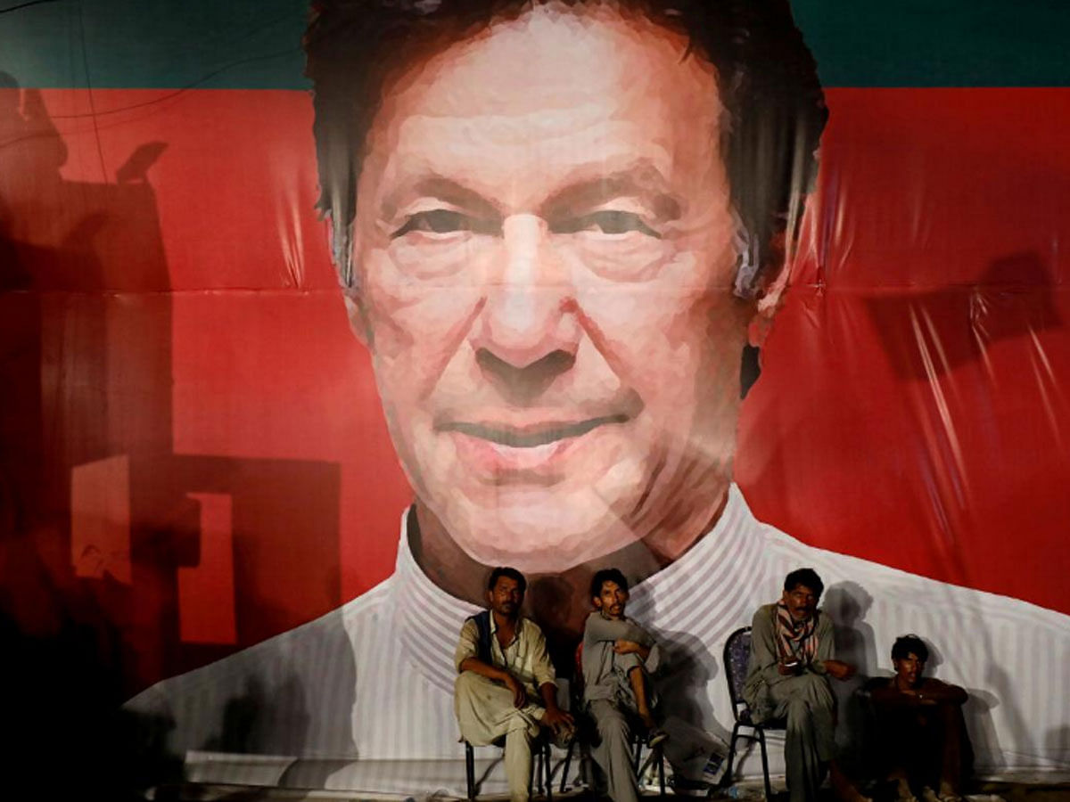 Labourers, who set up the venue, sit under a wall with a billboard displaying a photo of Imran Khan, chairman of the Pakistan Tehreek-e-Insaf (PTI), political party, as they listen to him during a campaign rally ahead of general elections in Karachi, Pakistan on 22 July 2018. -- Reuters