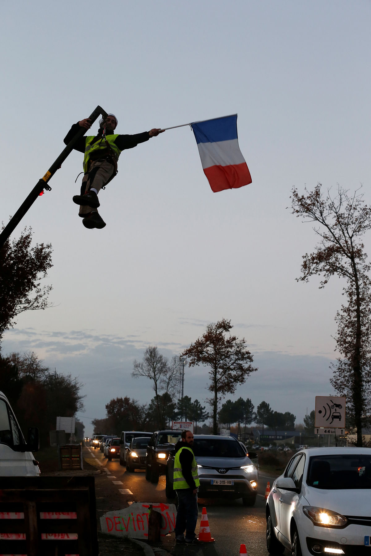 A protester wearing a yellow vest, the symbol of a French drivers` protest against higher diesel fuel prices, holds a French flag as he hangs by a crane at a roundabout occupied by yellow vests protesters in Cissac-Medoc, France, 5 December 2018. Photo: Reuters