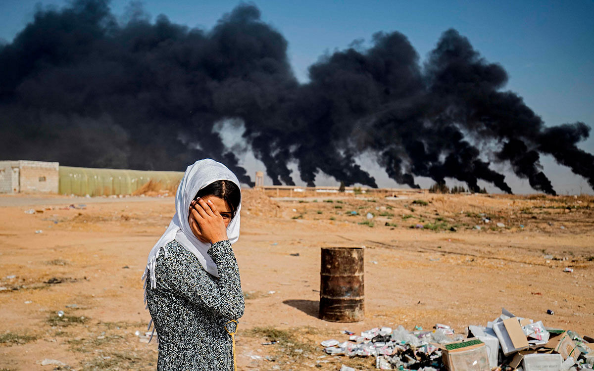 A woman covers her face as she stands along the side of a road on the outskirts of the town of Tal Tamr near the Syrian Kurdish town of Ras al-Ain along the border with Turkey in the northeastern Hassakeh province on 16 October 2019, with the smoke plumes of tire fires billowing in the background to decrease visibility for Turkish warplanes that are part of operation `Peace Spring`. Photo: AFP